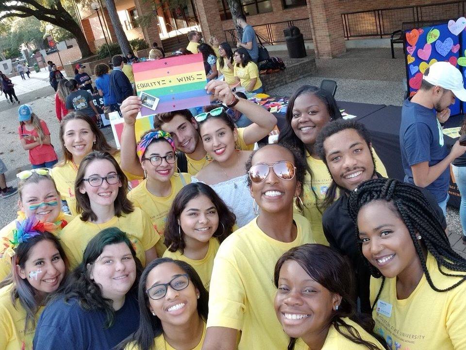 Students pose in matching yellow shirts with a pride flag sign being held in the background.